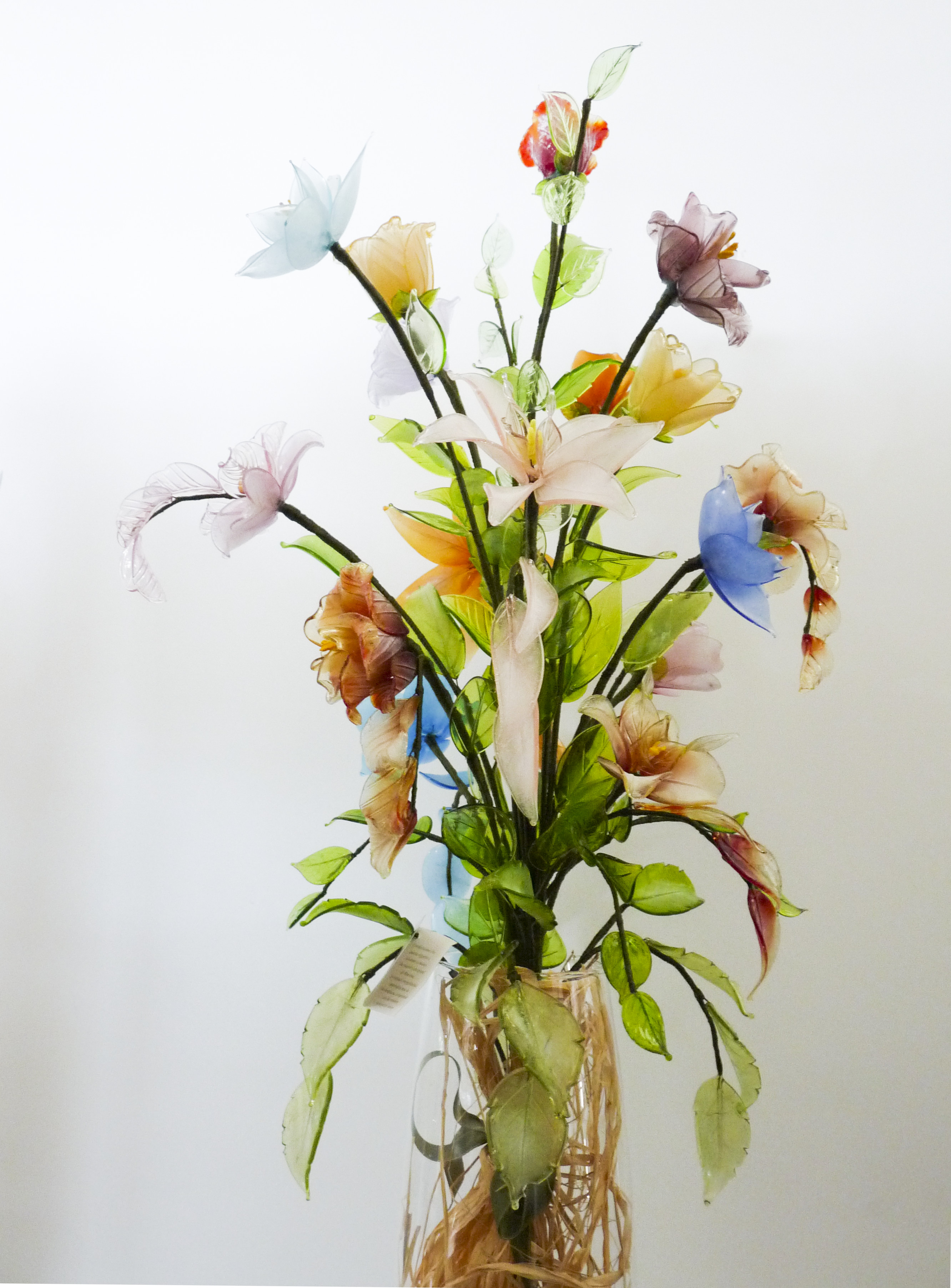 Vaso composizione di fiori in vetro, di Murano, assortiti per forma e dimensione, appoggiati su un vaso trasparente, immagine in primo piano.