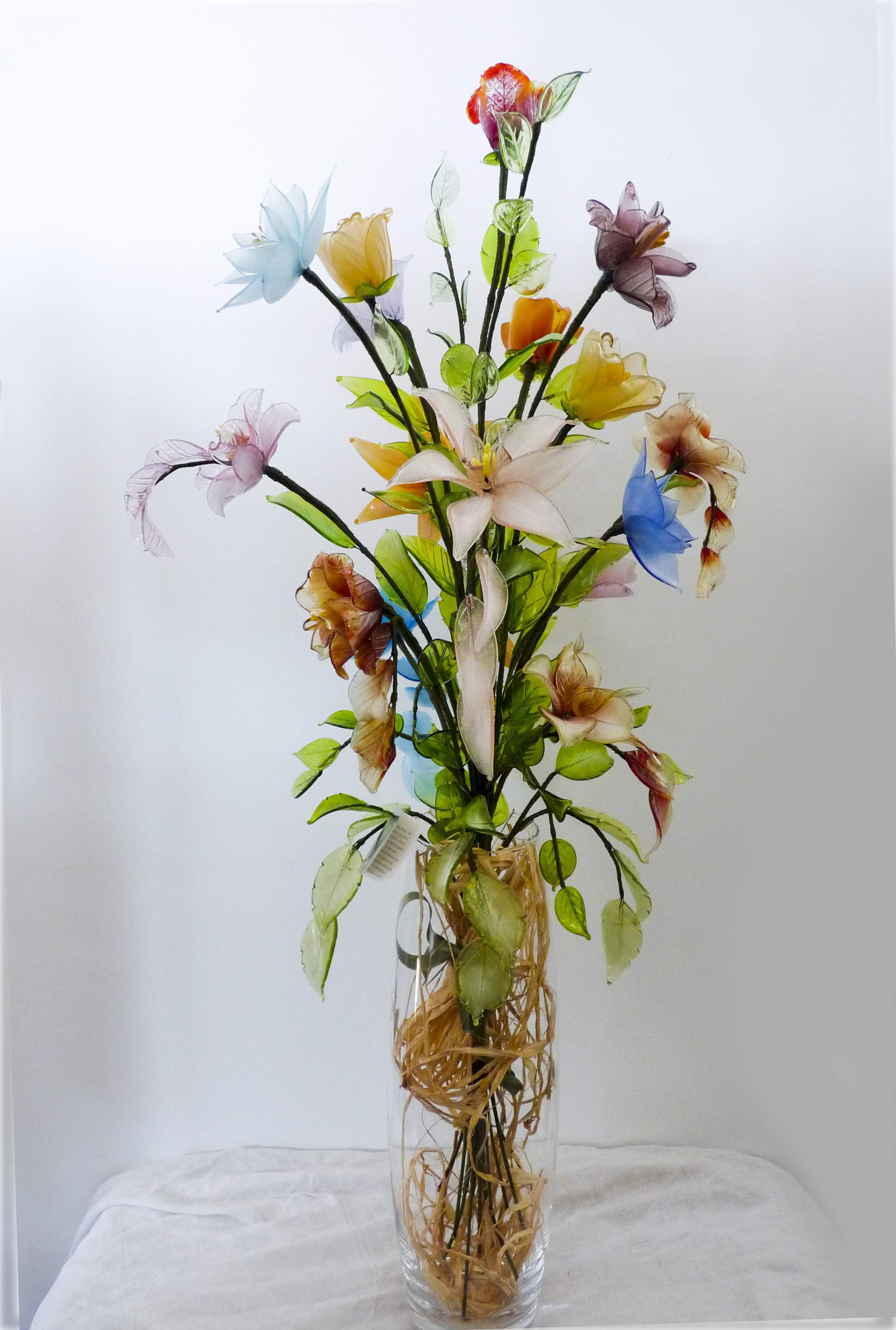 Vaso composizione di fiori in vetro, 
di Murano assortiti per forma e dimensione, appoggiati su un vaso trasparente.